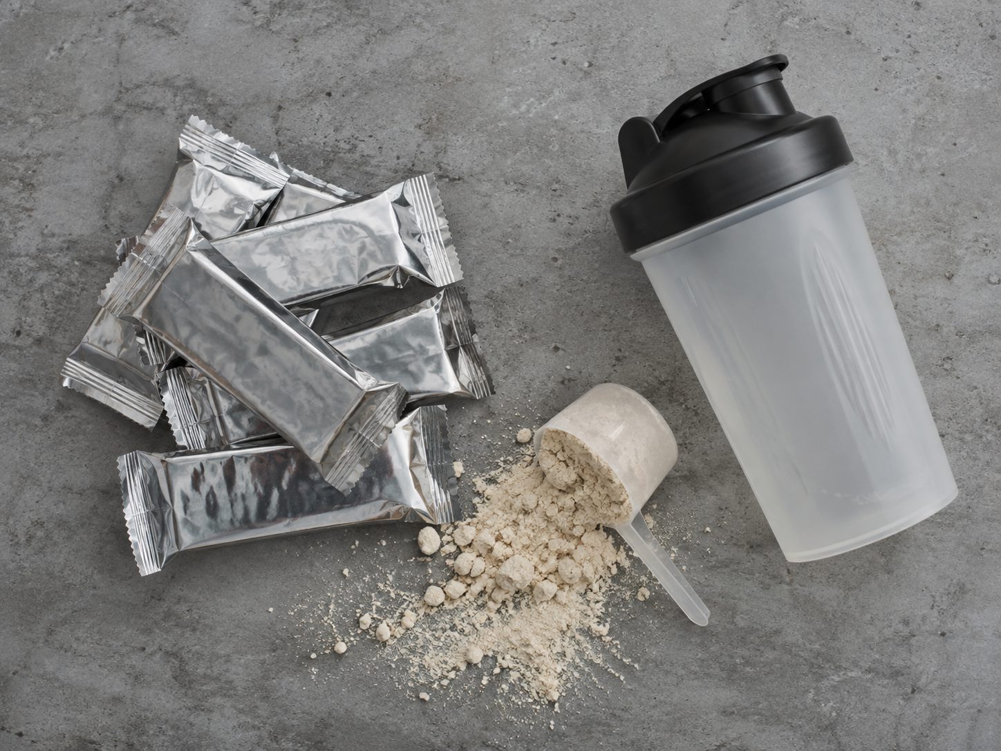 En haug proteinbarer i blank plast, en skje proteinpulver og en shaker på en grå benk.